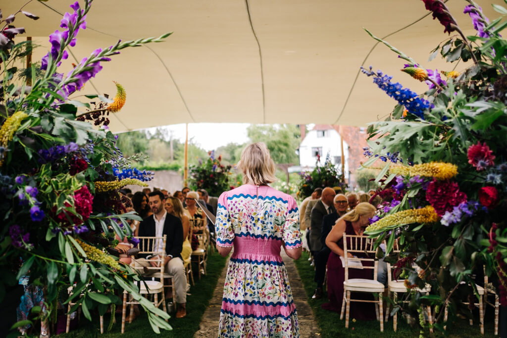 luxury marquee stretch tent ceremony at a private home in Kent with celebrant waiting for couple to come up the aisle surrounded by bold and bright flower urns
