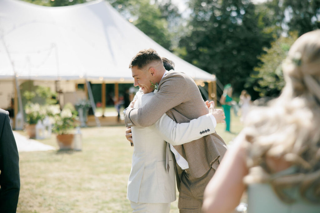 Groom and Best Man hugging during a drinks reception with a sail cloth marquee in the background at a wedding.