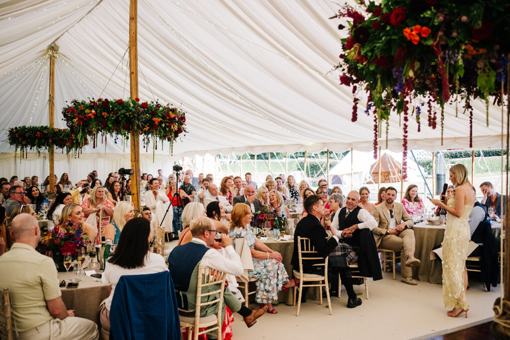 Maid of Honour giving a speech to 200 guests in a traditional pole tent with 3 abundant floral chandeliers around the poles with bright and bold florals.