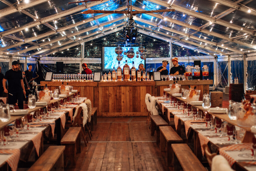 Clear roof marquee set up for a private apres ski themed winter party with a rustic wooden bar with large format bottles of Rose, rustic tables dressed with sheep skin blankets, vintage skis and disco balls hanging from the ceiling. 