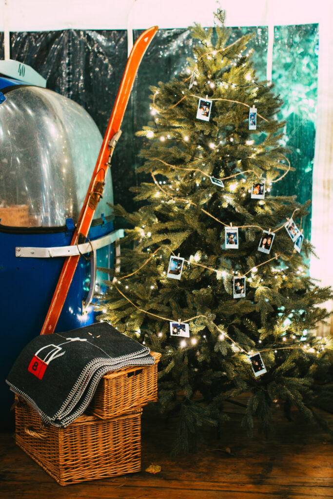 Christmas tree decorated with fairy lights and polaroid pictures of guests at a party next to a ski gondola prop, blankets and skis